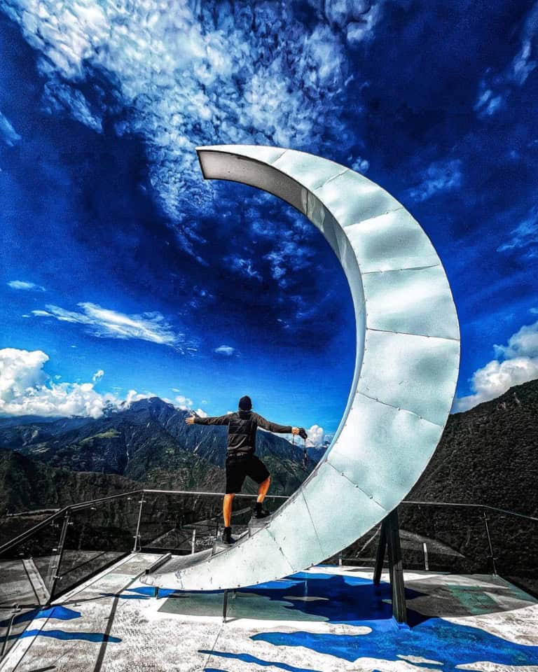 Person posing on metallic crescent sculpture overlooking mountains